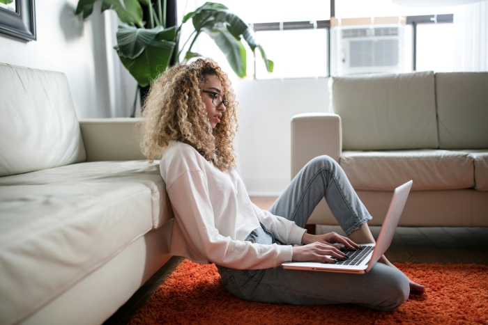 A person sits on the floor leaning against a sofa, doing research on their laptop.