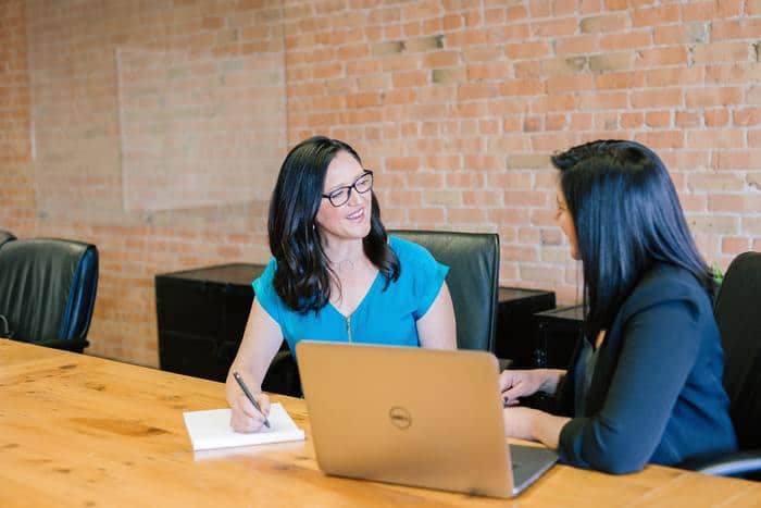 Two women in a meeting. They are talking to each other and there is a laptop and notepad on the desk.