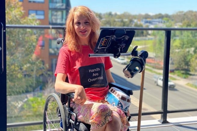 Gretta sitting in a wheelchair on a sunny balcony overlooking a tree-lined street.
