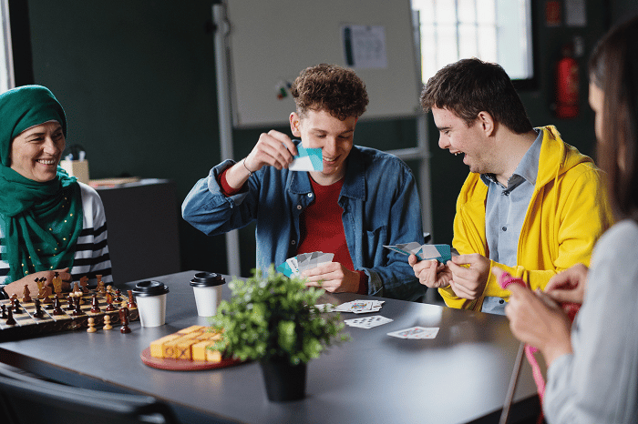 Four people sitting at a table playing cards. A chess board, takeaway coffee cups, and a plant are placed on the table.