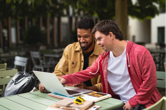 A person with disability works with their support worker at a laptop outside a cafe.