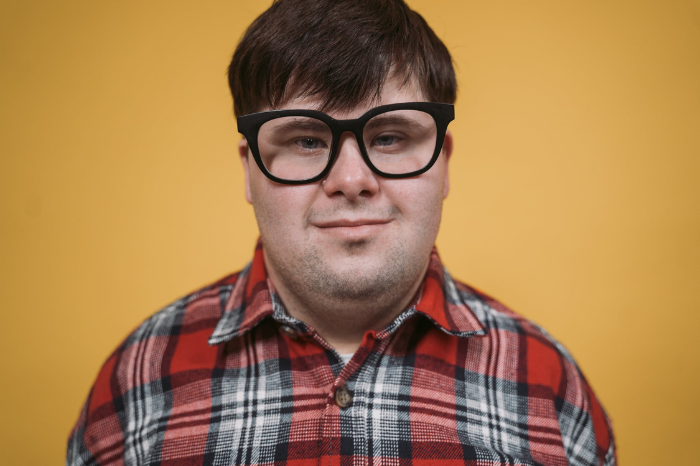 A young man wearing glasses and a plaid shirt stares confidently.