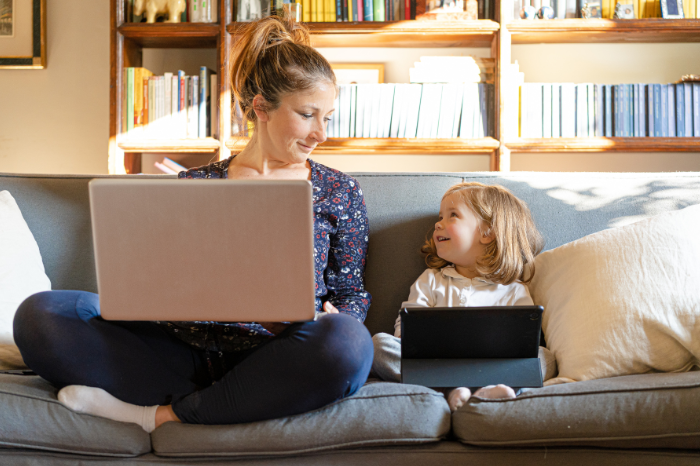 A mum and her daughter sit next to each other on a sofa. Both have laptops.