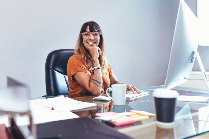 A smiling woman with glasses sits at her desk typing a report.