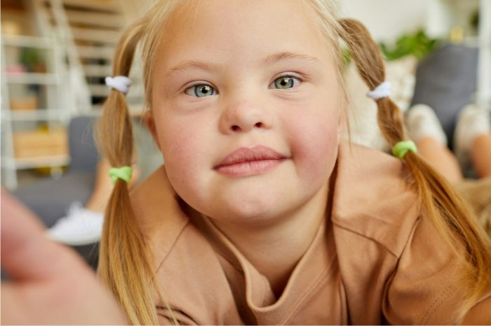 A close up of a young girl with blonde hair wearing pigtails and a tan-coloured top.